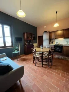 Woonkeuken met grijze bank, houten bruine eettafel en houten stoelen, er hangt een zwarte tv aan de wand